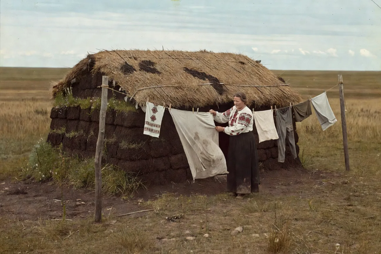 Authentic Ukrainian-Canadian sod house on the Manitoba prairie circa 1900, woman in traditional embroidered blouse hanging laundry, vast empty horizon