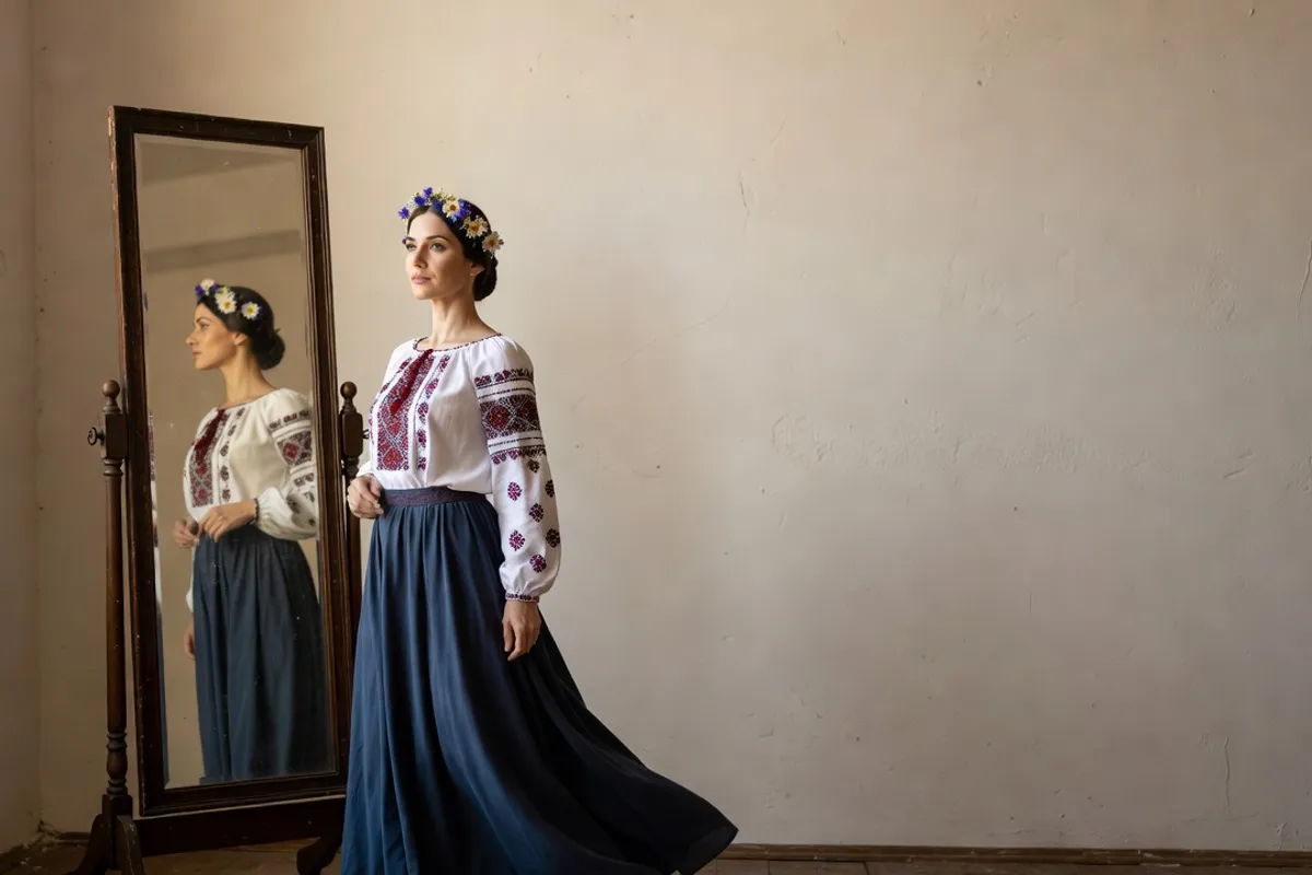 A dancer in a Ukrainian blouse and flower wreath standing beside a dressing mirror backstage.