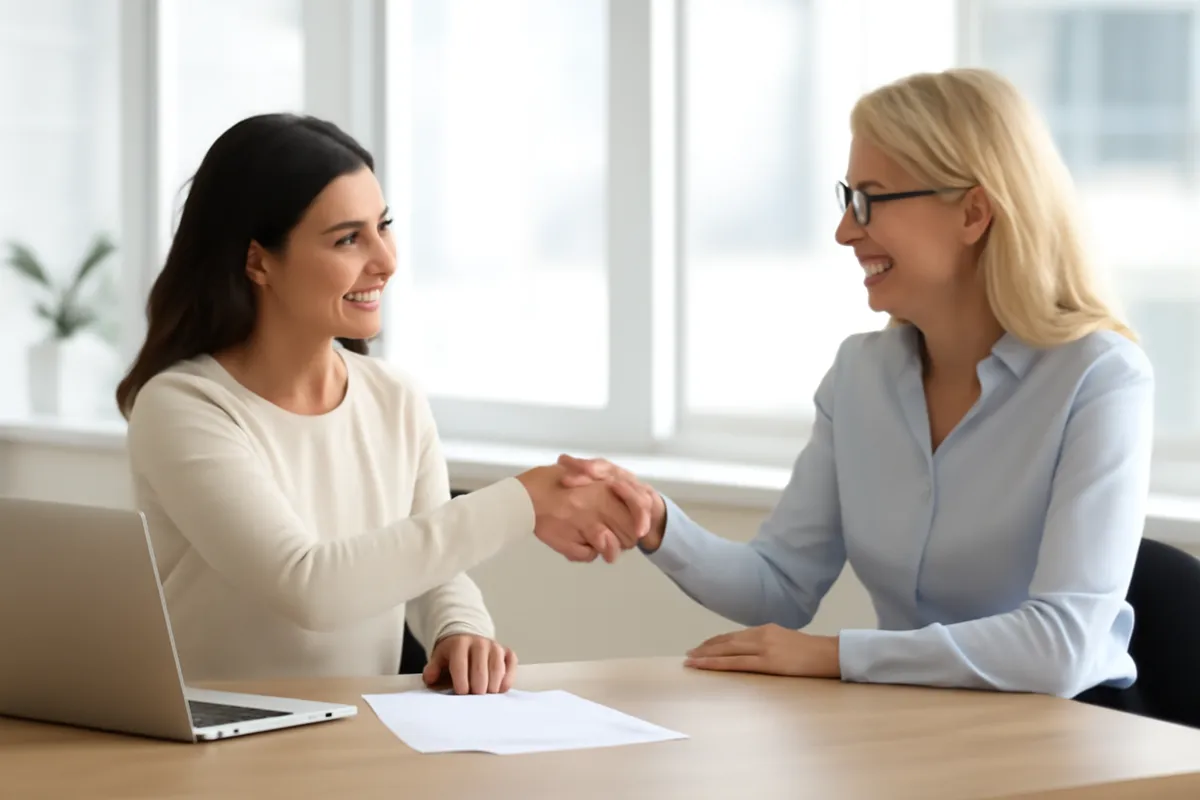 Two women shaking hands across a table in a bright office.
